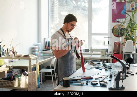 Junger, ernsthafter Kunstleder in Arbeitskleidung, der in der Werkstatt am Tisch steht und vor dem Schneiden roten Ledergürtel misst Stockfoto