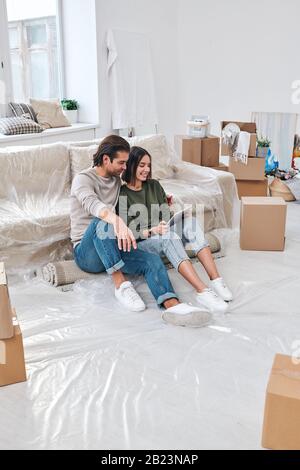 Junges amouröses Paar mit Touchpad, das auf gerolltem Teppich auf der neuen Couch sitzt, während die Suche nach einem Möbelangebot online ist Stockfoto