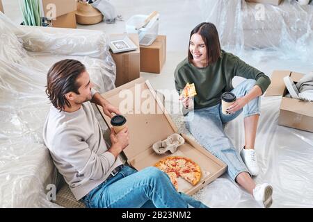 Fröhliche junge Frau mit einem Stück Pizza und einem Glas Kaffee, die mit ihrem Mann beim Mittagessen auf dem Boden des Wohnzimmers spricht Stockfoto