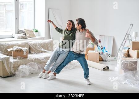 Junge lachende Frauen in der Opferkleidung, die sich an ihren Mann lehnt, während beide im Wohnzimmer tanzen, nachdem sie in eine neue Wohnung gebracht wurden Stockfoto