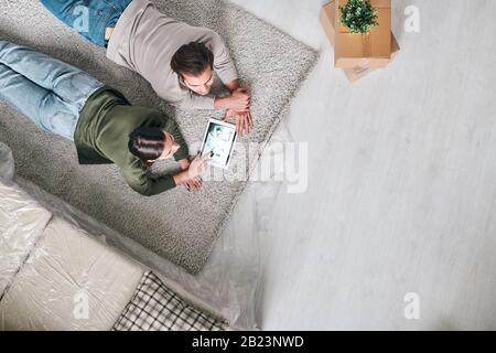 Draufsicht über junge, erholsame Paare, die auf dem Teppich liegen und durch Online-Werbung für neue Immobilien auf dem Touchpad scrollen Stockfoto