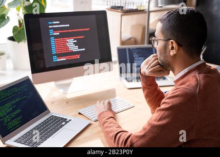 Junger, nachdenklicher Programmierer, der am Arbeitsplatz sitzt und während der Analyse Informationen auf dem Computerbildschirm betrachtet Stockfoto