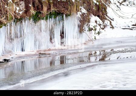 Eiszapfen, die an der Unterseite eines Flussufers hängen, spiegeln sich im letzten nicht gefrorenen Teil des Flusses wider. Stockfoto