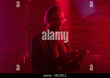 Junger seriöser Programmierer in Kopfhörern, der mobile Geräte verwendet, während er neue Software in roten Lichtern vorstellt Stockfoto