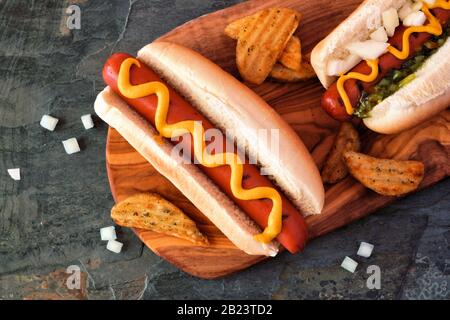 Heiße Hunde mit Belägen und Kartoffelkeilen auf Holzbrett. Nahaufnahme, Draufsicht. Stockfoto