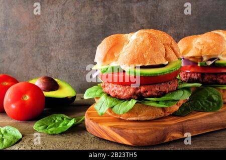 Fleischlose Burger mit Avocado, Tomate und Spinat auf Pflanzenbatterbasis auf einem Servierbrett vor dunklem Hintergrund Stockfoto