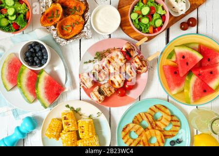 Sommer BBQ oder Picknick-Food-Konzept. Auswahl an Früchten, Salat, gegrilltem Fleisch und Kartoffeln. Tisch-Szene in der Draufsicht über einen weißen Holzhintergrund. Stockfoto