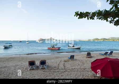 Schöner heller Tag am Strand, leere Strandstühle, ein einzelner Sonnenschirm und Fischerboote in der Nähe des Ufers am Reduit Beach, Gros Islet, Saint Lucia Stockfoto