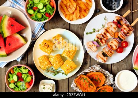 Sommer BBQ oder Picknick-Food-Konzept. Auswahl an Früchten, Salat, gegrilltem Fleisch und Kartoffeln. Überhängende Tisch-Szene auf rustikalem Holzhintergrund. Stockfoto