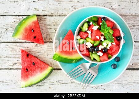 Wassermelonsalat mit Gurke, Blaubeeren und Fetakäse. Tisch-Szene in der Draufsicht auf weißem Holzhintergrund. Stockfoto