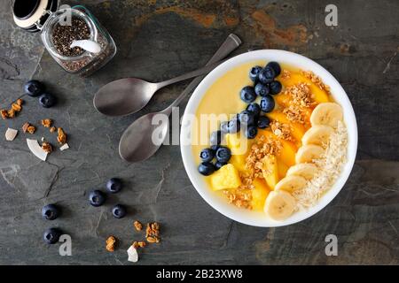 Gesunde Ananas, Mango-Smoothie-Schüssel mit Kokosnuss, Bananen, Blaubeeren und Granola. Über der Ansicht auf dunklem Hintergrund. Stockfoto
