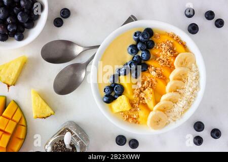 Gesunde Ananas, Mango-Smoothie-Schüssel mit Kokosnuss, Bananen, Blaubeeren und Granola. Szene in der Draufsicht auf hellem Hintergrund. Stockfoto