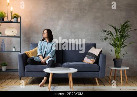 Junge Frau in Brille wütend auf das Telefon sitzend auf der Couch. Junge Geschäftsfrau. Stockfoto