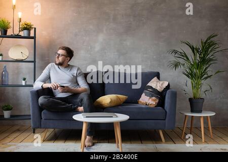 Junger Mann in Brille, wütend, am Telefon sitzend auf der Couch zu sprechen. Junger Geschäftsmann. Stockfoto