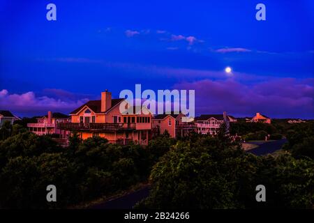 Erste Strahlen der Morgendämmerung leuchten auf den Ferienhäusern an der Küste gegen einen verblassenden Nachthimmel und Mond. Stockfoto