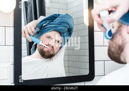 Der schlafende Mann mit einem Handtuch auf dem Kopf bürstet Zähne mit einer elektrischen Bürste und sieht mit Gleichgültigkeit auf seine Spiegelung im Spiegel aus. Stockfoto