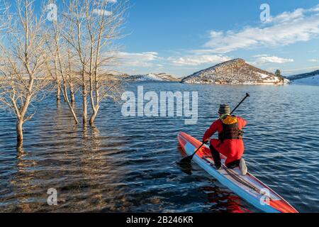 Ein männlicher Paddler, der einen Trockenanzug, eine Schwimmweste und eine Sicherheitsleash trägt, beginnt mit dem Training auf einem langen Rennstativ am Paddleboard auf einem Bergsee in Colorado Stockfoto