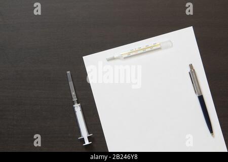 Vorlage für weißes Papier mit Stift, Thermometer und Spritze für die Injektion auf dunklem Holzhintergrund in Wenge-Farbe. Die Bereitstellung medizinischer und pharmakologischer Dienstleistungen. Mit leerem Leerzeichen für Text. Stockfoto