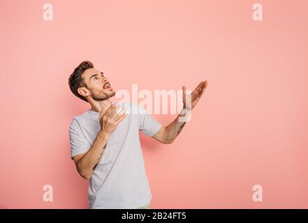Staunender junger Mann, der aufblickt, während er mit offenen Armen auf pinkfarbenem Hintergrund steht Stockfoto