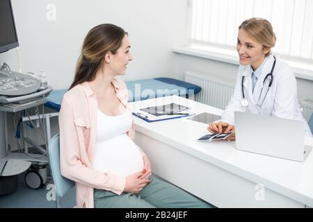 Lächelnder Arzt, der der jungen schwangeren Frau in der Klinik einen Ultraschall-Scan zeigt Stockfoto