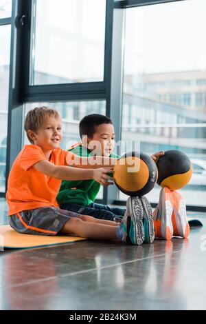 Multikulturelle Kinder setzen Bälle auf Zehen und sitzen auf Fitnessmatte im Fitnessstudio Stockfoto