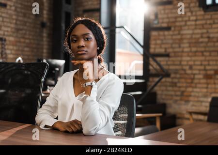 Attraktive, nachdenkliche afroamerikanische Geschäftsfrau, die am Schreibtisch sitzt und die Kamera betrachtet Stockfoto