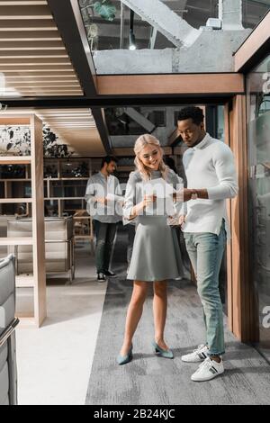 Ein ernsthafter afroamerikanischer Geschäftsmann, der mit einem lächelnden Kollegen ein Dokument betrachtet, während multikulturelle Freunde vor dem Hintergrund stehen Stockfoto