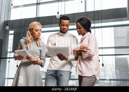 Junge Geschäftsfrau, die digitales Tablet hält, während sie in der Nähe afroamerikanischer Kollegen steht, die Dokumente betrachten Stockfoto