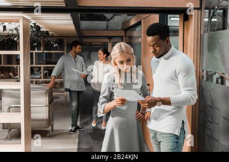 Ein gutaussehender afroamerikanischer Geschäftsmann, der mit einem hübschen Kollegen Dokumente betrachtet, während multikulturelle Freunde auf dem Hintergrund stehen Stockfoto
