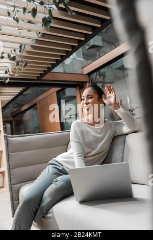 Selektive Fokussierung der glücklichen asiatischen Geschäftsfrau, die beim Sitzen auf der Couch in der Nähe eines Laptops die Hand winkt Stockfoto