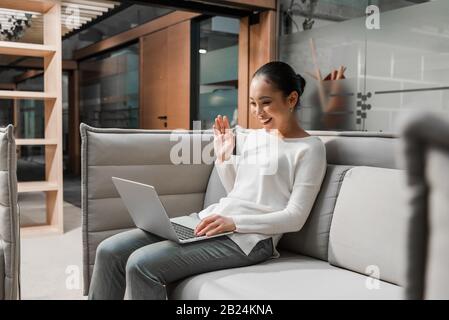 Glückliche asiatische Geschäftsfrau winkt beim Sitzen auf der Couch und beim Video-Chat auf dem Laptop Hand Stockfoto
