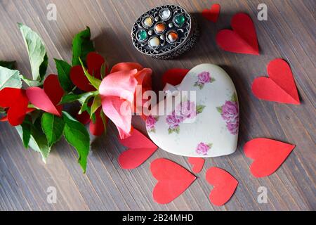 Herz von Porzelain mit Blumenmuster auf dem Holztexturtisch, rosa Rose und vielen roten Papierherzen. Flaches Lay. Geschenk und Begrüßung für Valentines D. Stockfoto