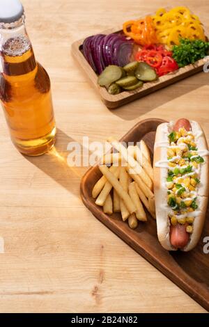 Köstlicher heißer Hund in der Nähe von Bord mit Gemüse in Scheiben, Bier und pommes frites auf Holztisch Stockfoto