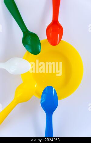 Mehrfarbige Kunststoff-Löffel, die sich um die gelbe Platte auf dem weißen Hintergrund befinden. Flaches Lay. Draufsicht. Mehrfarbiges Konzept. Stockfoto