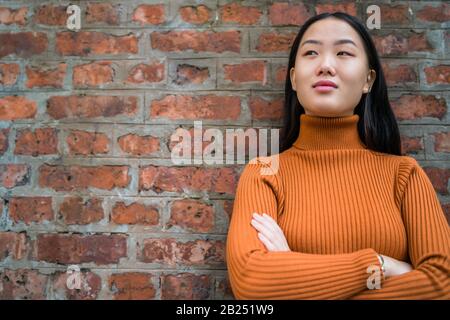 Nahaufnahme der jungen schönen asiatischen Frau, die selbstbewusst aussieht und vor Backstein-Wandhintergrund steht. Stockfoto