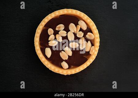 Süßes Tartlet mit Schokolade und Erdnüssen obenauf. Mürbeteig mit Nüssen und Schokoladenfondant auf schwarzem Grund. Stockfoto