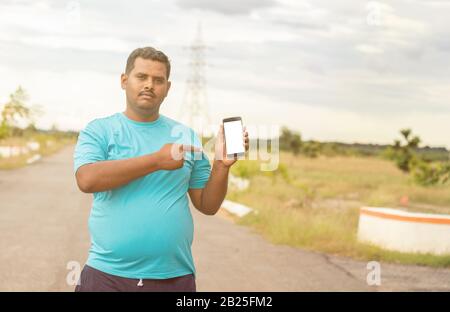 Junger Mann, der ein Joggingkleid trägt, das den Smartphone-Bildschirm zeigt, indem er mit der Hand und dem Finger auf den Park zeigt - Konzept der empfohlenen Fitness-Tracker-App auf Stockfoto