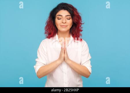 Achtsamkeitspraxis. Porträt einer friedlichen Hüftfrau mit ausgefallenen roten Haaren, die die Hände in namaste oder Geste hält und mit geschlossenen Augen meditiert. Stockfoto