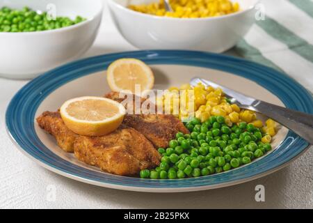 Goldbauch frittiertes Fischfilet, serviert mit grünen Erbsen, süßem Mais und Zitronenscheiben auf einem Teller auf weißem Grund Stockfoto