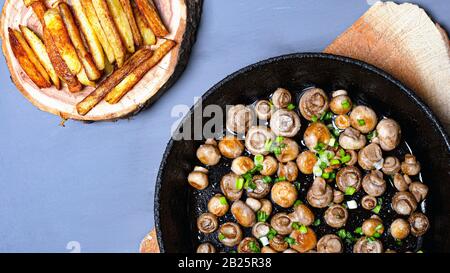 Gebratene Champignonpilze in einer gusseisernen Pfanne mit grünen Zwiebeln und Pommes frites auf einem Holzständer auf grauem Hintergrund Draufsicht. Stockfoto