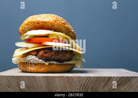 Saftiger Burger mit Häckchen, Käse und Gemüse auf grauem Grund mit Kopierraum, Doppel-Cheesebburger auf Holzbrett. Stockfoto