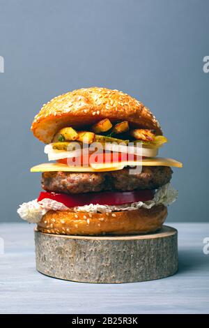 Köstlicher Burger mit einem großen Fleischesseln mit Gemüse, Käse und Pommes auf einem Holzwaldständer auf grauem Hintergrund. Stockfoto