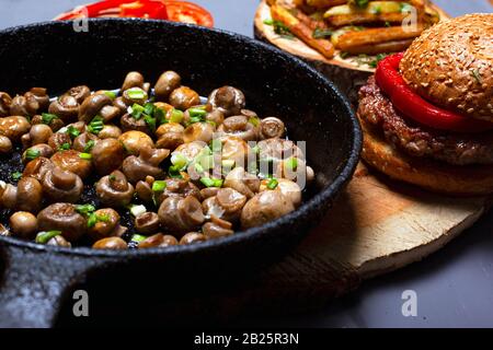 Golden gebratene Champignon in einer schwarzen Pfanne und einem saftigen Burger, hausgemachter köstlicher Vorspeise, fetthaltige Speisen auf grauem Grund. Stockfoto