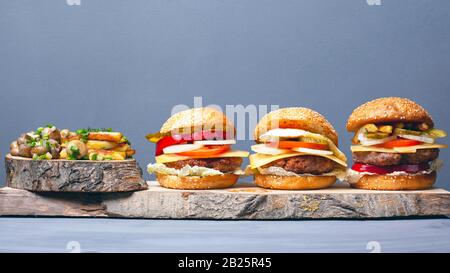 Drei leckere Hamburger mit Pommes und Champignons, die mit grünen Zwiebeln auf grauen Holzküsten bestreut werden. Herzhaftes Mittagessen. Stockfoto