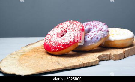 Drei verschiedene glasierte Krapfen in verschiedenen Farben auf einer Holzplanke auf grauem Grund. Stockfoto