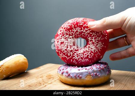 Weibliche Hand hält süßes Gebäck, Donuts mit roter und violetter Glasur auf einem Holzständer auf grauem Hintergrund in Nahaufnahme. Stockfoto