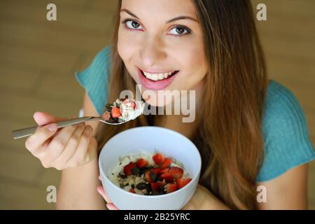 Nahaufnahme von oben der schönen jungen Frau lächelnd und beim Essen von Skyr mit Müsli Müsli Obst und Samen zu Hause, Fokus auf Modellaugen, Innenbild. Stockfoto