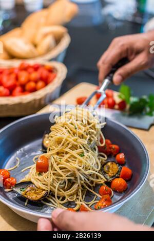 Nahaufnahme eines Chefkochs, der Spaghetti Vongole mit roma-tomaten und Muscheln in einer Pfanne zubereitet - mit Messer und Schneidebrett mit Tomaten und Brot im Rücken Stockfoto