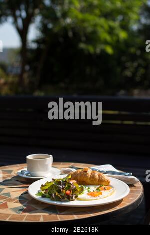 Frühstück mit Croissant, Sunny Side-up-Eiern, Salat und Kaffee auf einem runden Tisch im Garten Stockfoto