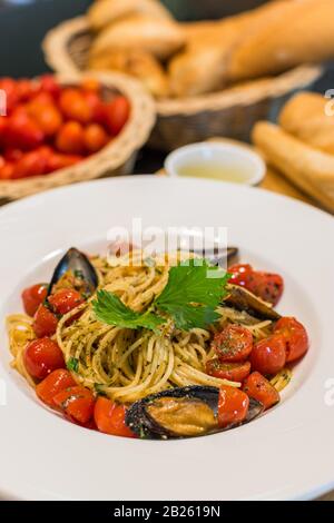 Nahaufnahme von Spaghetti Vongole mit roma-tomaten und Muscheln serviert und auf einem Teller mit Tomaten, Brot und Olivenöl im Hintergrund dekoriert Stockfoto